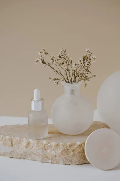 Aesthetic minimalist beauty care therapy concept. Organic serum oil cosmetics bottle on stone with flowers against neutral beige background. Body skin, face treatment product composition