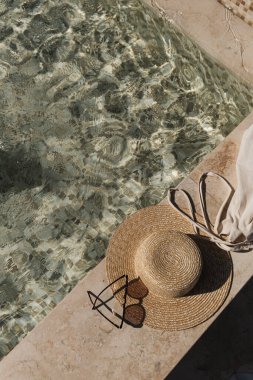 Flat lay of sunglasses and straw hat on marble swimming pool side with clear blue water with waves sunlight shadow reflections. Minimal fashion aesthetic summer vacation top view creative background clipart