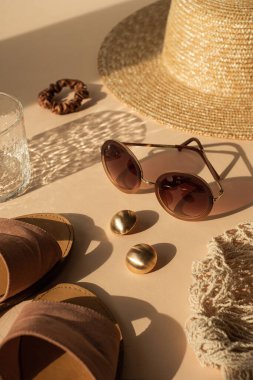 Parlak güneş ışığı gölgelerinde kristal camlı lüks bohem estetik kompozisyonu. Güneş gözlüğü, kadın hasır şapka, sicim torbası, altın küpeler, tarafsız bej arka planda terlikler.