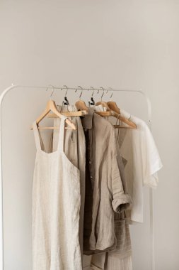 Minimum estetik moda kıyafetleri konsepti. Tarafsız bej çamaşır, kadın bluzları, elbiseler ve beyaz arka planda asılı tişörtler. Moda blogu, web sitesi, sosyal medya