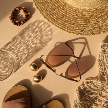 Parlak güneş ışığı gölgelerinde kristal camlı lüks bohem estetik kompozisyonu. Güneş gözlüğü, kadın hasır şapkası, sicim torbası, tarafsız bej arka planda altın küpeler. Düz yatış, üst görünüm