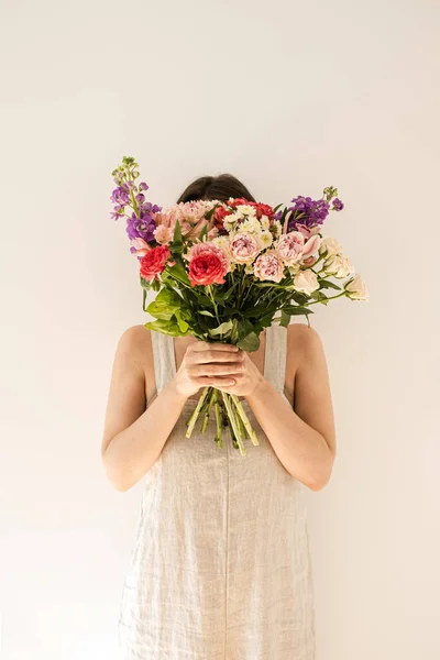 ベージュ洗浄リネンのサンドレスの若い美しい女性は白い壁に対してカラフルなバラの花の花束を保持します 美しい休日のお祝いの花組成 — ストック写真