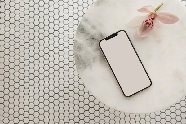 Blank clipping path screen mobile phone, pink flower on marble stone table on mosaic tile background. Aesthetic template with mockup space. Flat lay, top view