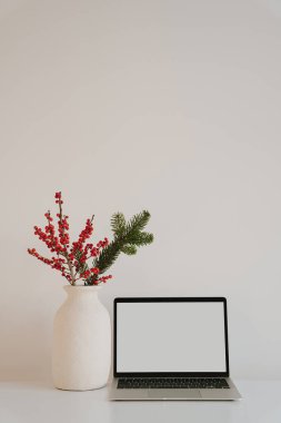 Boş ekranlı dizüstü bilgisayarı olan basit bir ofis ve beyaz bir masanın üzerinde kırmızı böğürtlen ve çam dallarıyla tatil temalı vazosu. Minimalist ve konforlu çalışma alanı dekoru