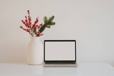 Dizüstü bilgisayarı, kırmızı böğürtlen ve çam filizlerinden oluşan dekoratif vazosu olan minimalist Noel temalı çalışma alanı. Temiz ve estetik ofis kurulumu