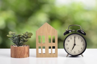 Çalar saati ve küçük bitkisi olan ahşap ev modeli. Gayrimenkul zamanlaması, emlak yatırım planlaması, konut piyasası fırsatı ve ev sahipliği kavramı.