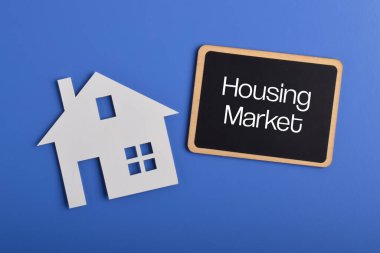 Konut piyasası kavramı. Mavi arkaplanda ev silueti ve yazı tahtası var. Emlak eğilimleri, emlak yatırımları, ev satın alma ve konut piyasası analizi.