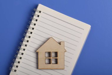 Mavi arka planda izole edilmiş boş bir defter üzerinde minyatür ahşap bir ev. Gayrimenkul planlaması, ipotek hesaplaması, ev arama veya mülk yönetimi kavramı.