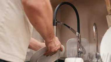 Erkek figürü şık bir mutfak lavabosunda bulaşık yıkamakla meşgul, ovma eylemi ve su akışını vurguluyor, yavaş yavaş göreve yakınlaşıyor