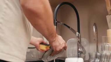 Erkek figürü, çağdaş bir mutfak lavabosunda bulaşık yıkamakla meşgul, su akışını ve bulaşık yıkama sürecini vurguluyor, kamera detaylar için yakınlaşıyor