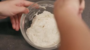 Eller, cam bir kâsede kalın hamuru aktif olarak harmanlıyor, kremalı dokuları ve karıştırma tekniğini vurguluyor, kamera büyüleyici bir yemek pişirme deneyimi için yakınlaştırıyor.