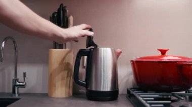 Paslanmaz çelik çaydanlık, renkli yemek takımları ve çatal bıçaklarla çevrili çağdaş bir mutfağın kaynama sürecini ön plana çıkaran bir el tarafından açılır.