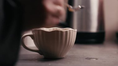 El, mutfak tezgahındaki seramik bardağa şeker karıştırıyor. Ilık bir içeceğin hazırlığını vurguluyor.