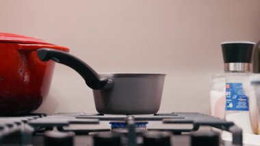 Mutfak sahnesinde, kırmızı bir tencere ve baharatla çevrili bir tencereye yumurtayı koyan bir el yemek pişirme eylemini, kamera tavalarını ve yakınlaşmalarını betimliyor.