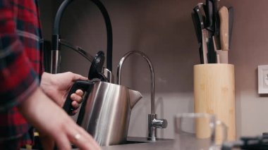 Ekose gömlekli erkek figürü, ısıtıcıdan cama sıcak su dökmek üzere. Mutfak aletlerini ve aletlerini vurguluyor ve davetkar bir atmosfer yaratıyor.