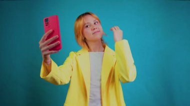 Elinde pembe bir akıllı telefon tutan gülümseyen genç bir kadın, poz verirken ve el sallarken selfie çekiyor ve video görüşmeleri yapıyor, sağlam bir deniz mavisi stüdyosuna karşı mutluluk ve güveni ifade ediyor.