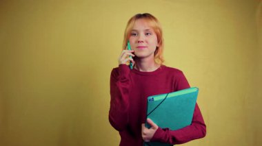 Elinde dosya tutarken ciddi bir telefon görüşmesi yapan profesyonel bir genç kadın. Endişeli ve düşünceli olmaktan mutlu ve başarılı olmaya kadar bir dizi duyguyu sarı bir arkaplanda sergilemek.