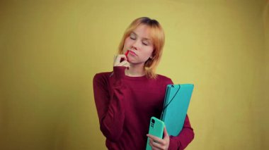 Bir klasör ve akıllı telefon tutan yaratıcı genç öğrenci gülümsemeden önce çenesini bir kalemle hafifçe vuruyor, ilham veriyor ve saf sarı arka planda bir çözüm buluyor.