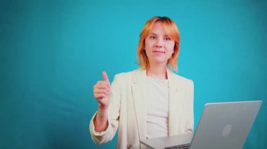 Elinde dizüstü bilgisayarla gezen, gülümseyen, olumlu bir işaret veren, stüdyoda mavi bir arka plandan memnun olduğunu belirten, olumlu genç bir kadın.
