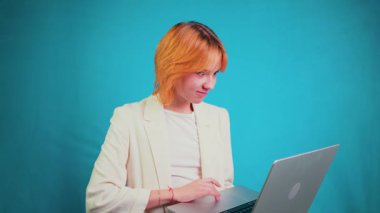 Portakal rengi saçlı neşeli genç bir profesyonel, video görüşmesine katılıyor, aktif olarak iletişim kuruyor, el sallıyor ve dizüstü bilgisayarını mavi arka planda kullanırken çeşitli duyguları ifade ediyor.