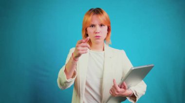 Kızıl saçlı, kendine güvenen genç bir kadın kameraya ciddi bir ifadeyle bakıyor. Elinde dizüstü bilgisayarla mavi bir zemine karşı durarak bir seçim yapıyor.