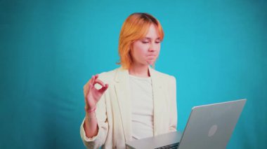 Kendine güvenen genç bir kadın elinde dizüstü bilgisayarla iyi bir jest yapıyor, gülümsüyor ve bir projeyi onaylarken sağlam turkuaz mavisi bir arka planda tek başına duruyor.