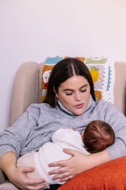 Sevgi dolu anne, yeni doğmuş bebeği rahat bir sandalyede emzirir, bebeğe yakın durur ve evde şefkatli bir bağ, rahatlık, rahatlama ve annelik duygusunu paylaşır.