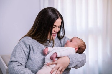 Genç anne, yeni doğmuş bebeğini kucaklarken sıcak bir şekilde gülümsüyor. Evdeki anne bağını, şefkatini ve neşeli bağını yansıtan şefkatli bir anı paylaşıyor.