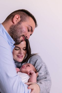 Yeni anne-babalar bebeklerini kucağında tutarlar. Sevgi ve bağlantının samimi bir anını paylaşırlar. Beyaz bir arka plana karşı ailenin ve ebeveynliğin neşe ve şefkatini somutlaştırırlar.