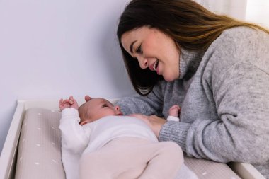 Anne gülümsüyor ve yeni doğmuş bebeğiyle bağ kuruyor. Değişen bir masada yatıyor. Şefkati, aile sevgisini ve erken annelik anlarını simgeliyor.