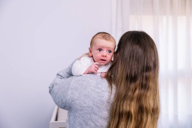 Genç bir kadın, bebeğini nazikçe omzunda tutuyor. Parlak bir ev ortamında sevgisini ve bakımını ifade ediyor. Erken yaşta çocuğuyla bağ kuruyor.