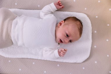 Yeni doğmuş erkek bebek bej benekli bez bir karyolada sırt üstü uzanıyor, sakin, rahat bir ifadeyle yan yana bakıyor, pamuk havluya sarılmış, güvende ve huzurlu hissediyor.