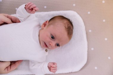 Yumuşak beyaz bir pedin üzerinde uzanan yeni doğmuş bebek, gözleri açık ve meraklı ebeveynler küçük çocuğu nazikçe destekliyor ve rahatlatıyor, şefkati ve özeni çağrıştırıyor.