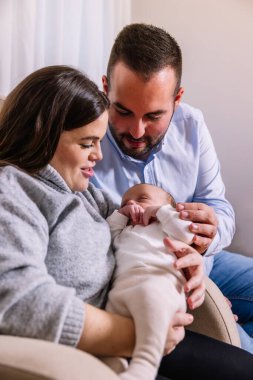 Mutlu ebeveynler, uyuyan yeni doğan bebeklerini kollarında tutarak yeni aile bağlarının neşe ve şefkatini kucaklayarak rahat bir ev ortamında birbirlerine sarılırlar.