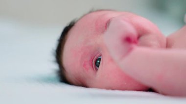 Yeni doğmuş bebek, yumuşak bir battaniyenin üzerinde uzanırken komik yüz ifadeleri ve ağız hareketleri yapıyor. Merakın ve reflekslerinin erken belirtilerini gösteriyor. Güzel mavi gözleri odaya bakıyor.