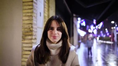 Uzun saçlı neşeli genç bir kadın kameraya bakıyor ve yağmurlu bir şehir sokağında gülüyor. Bulanık şehir ışıklarının arka planına karşı mutluluk ve neşe dolu büyüleyici bir portre.