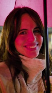 Güzel genç bir kadın, gece pembe bir şemsiyenin altında dururken mutlu bir şekilde gülümsüyor. Yüzü, canlı şehir ışıklarıyla aydınlanıyor ve elinde bir fincan kahve tutuyordu.