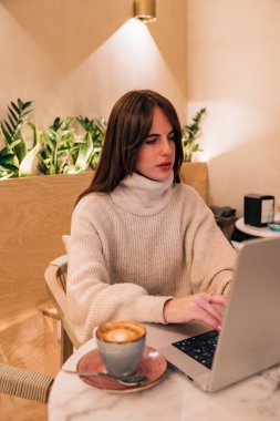 Dizüstü bilgisayarda yazan, kahvenin tadını çıkaran, ve sıcak ışıklandırma ve yeşil bitkilerle modern bir kafede uzaktan çalışan, üretkenliği somutlaştıran ve serbest yaşam tarzını kolaylaştıran bir kadın.