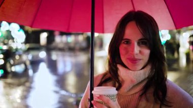 Yağmurlu bir gecede elinde kahve tutan ve kırmızı bir şemsiye altında gülümseyen neşeli bir kadının portresi. Bulanık sokak lambaları güzel bir arka plan oluşturuyor.
