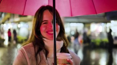 Güzel genç bir kadın kameraya gülümsüyor, elinde kırmızı bir şemsiye ve bir fincan kahve ile yağmurlu bir şehir caddesinde, arka planda renkli ışıklarla şehir atmosferinin tadını çıkarıyor.