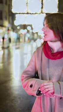 Güzel genç bir kadın gece bir şehir caddesinde kameraya bakıp gülümsüyor. Etrafı bulanık Noel ışıkları ve tatil sezonu boyunca ıslak kaldırımda yansımalarla çevrili.