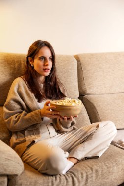 Kadın rahat bir kanepede oturup film seyrediyor ve ahşap bir kaseden lezzetli patlamış mısırlar yiyor, evinde rahat bir eğlence ve boş zaman akşamını kucaklıyor.