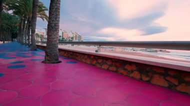 Güzel bir gün batımında, kumlu bir sahil ve şehir binalarının yanında palmiye ağaçlarıyla kaplı canlı pembe ve mavi fayanslı bir gezinti alanında yürüyen birinin alçak açılı görüntüsü.