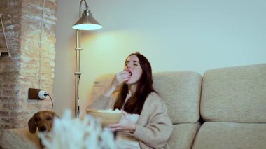 Genç bir kadın geceleri kanepede dinleniyor, bir kaseden patlamış mısır yiyor ve film izlerken dachshund 'unu okşuyor, oturma odasında rahat bir atmosfer yaratıyor.