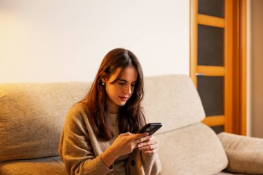 Evdeki bir koltukta rahat bir şekilde oturan genç bir kadın, cep telefonuna dalmış, iletişim, sosyal medya veya eğlence için online teknolojiyi bir anda kullanıyor.