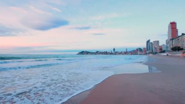 Kumlu sahilin güzel panoramik manzarası ve sakin deniz dalgaları Benidorm, İspanya 'nın kentsel ufuk çizgisi arka planda renkli bir gündoğumu sırasında modern apartman binalarını gözler önüne seriyor.