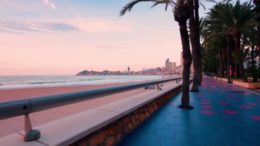 Benidorm, İspanya 'daki canlı Poniente plajı boyunca hareket eden kamera kumlu sahili, sakin denizi ve güzel, yumuşak, pembe ve mavi gün batımında görünen şehir silueti.