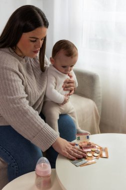 Anne bebeğini kucağında tutarken bir masanın üzerindeki euro banknotlarını ve sikkeleri dikkatle sayıyor, mali yönetimi, aile bütçesini ve ebeveynlik masraflarını temsil ediyor.