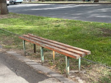 Yeşil çimlerin yanındaki kaldırımda boyası soyulan eski ahşap bir park bankında. Yıpranmış açık hava koltuğu zamanı, basitliği ve kentsel bozulmayı simgeliyor..