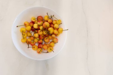 Beyaz mermer bir yüzey üzerinde sarı ve kırmızı kirazlarla dolu beyaz bir kasenin yakın çekim görüntüsü geniş kopya alanı ile düz görünüm